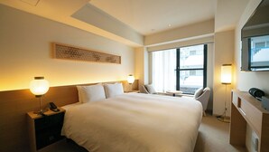 Quarto de hotel com uma cama grande, luminárias de cabeceira, uma escrivaninha, uma cadeira e uma janela com vista.