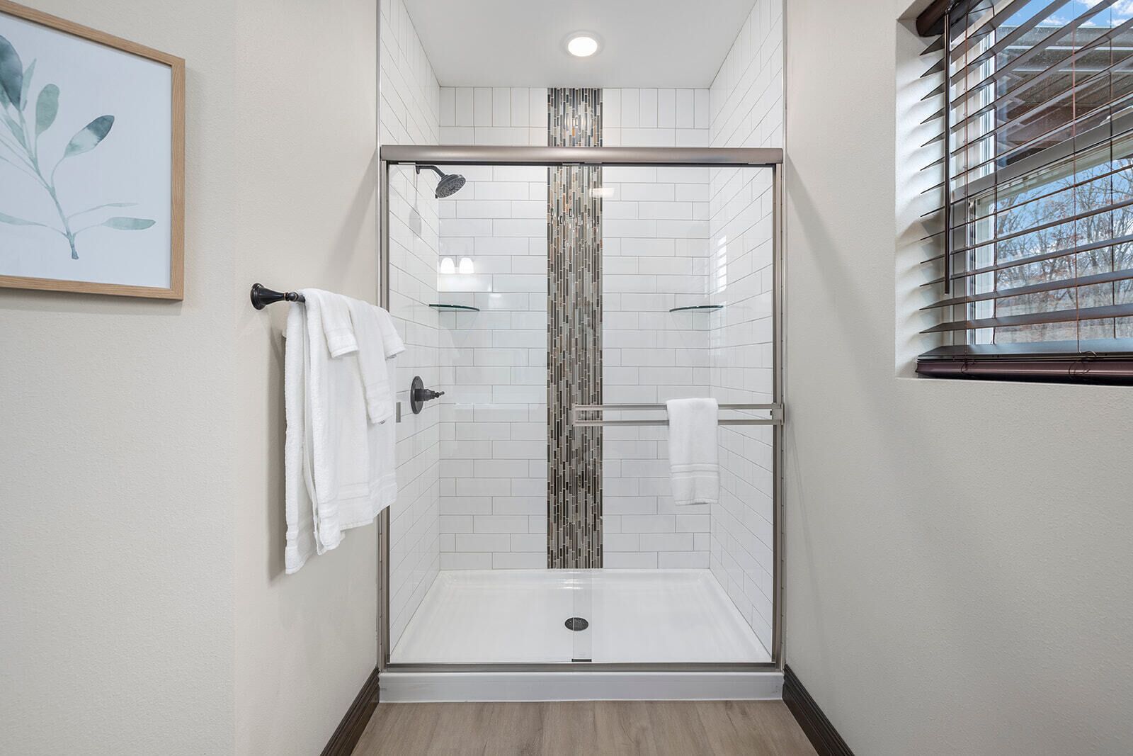 A glass-enclosed shower with white tiles, a framed plant on the wall, and a window with blinds.