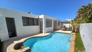 A swimming pool with a stone edge, a white building with barred windows, and a patio area with chairs and a table. - San Juan, Garden suite w/private POOL, outdoor kitchen/BBQ & peaceful (San Juan)