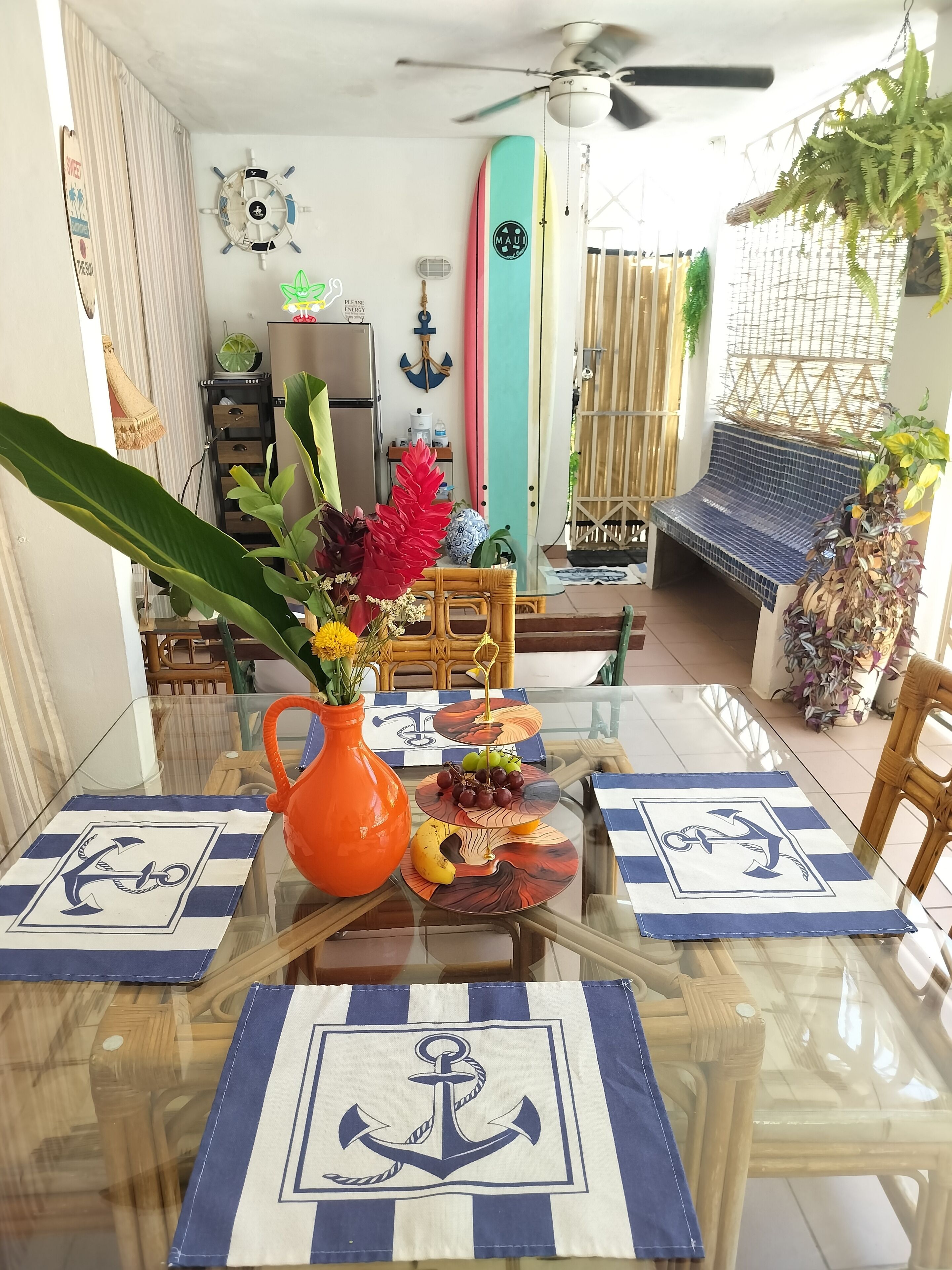 Un comedor con una mesa de vidrio, individuales a rayas azul y blanco, y un jarrón naranja central con flores.