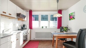 A compact kitchen with white cabinets, a stove, and a sink. There is a window with red curtains, a radiator below, and a table with a chair. - Ferienwohnung mit Ausblick (Dalaas Wald)