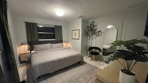 Een slaapkamer met een bed, bedlampen, een raam met gordijnen, een spiegel, een plant in pot en een dressoir.