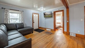 Un salon comprenant un canapé d’angle, un plancher en bois, un téléviseur fixé au mur et une porte donnant sur une autre pièce.