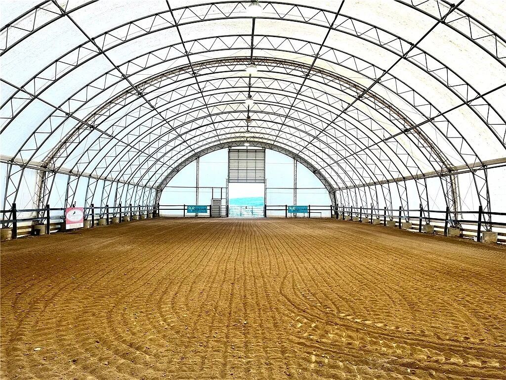 An indoor arena with a metal roof and sandy floor, featuring a central viewing area and railings.