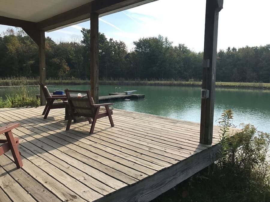 A wooden deck with two chairs and a table, overlooking a calm lake with a dock.