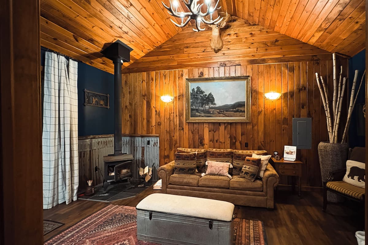 A cozy cabin interior with a wooden ceiling, a fireplace, a sofa with patterned cushions, and a rug.