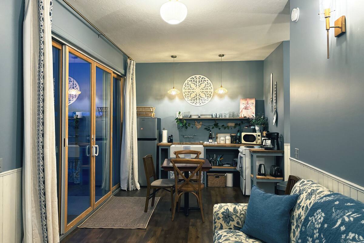 A compact kitchen with a refrigerator, microwave, and coffee maker, a dining area with a table and chairs, and a sofa with a patterned cushion.