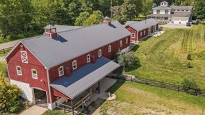 A large red barn with a covered area and a smaller adjacent building, surrounded by lush greenery and a hill. - Letchworth Farm Guesthouses (Nunda)
