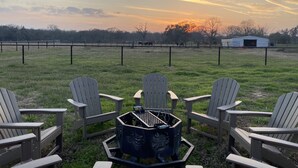 A grassy outdoor area with Adirondack chairs arranged around a portable grill, a fenced pasture in the background, and a barn to the right.