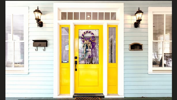 A house with a bright yellow front door, two windows with shutters, and two wall-mounted lanterns. - Welcome to House on Hallett.  A historic home less than a mile from downtown (Mobile)