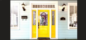 Una casa amb una porta principal de color groc brillant, dues finestres amb persianes i dos fanals penjats a la paret.