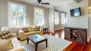 A living room with a sofa, coffee table, and TV, featuring large windows and a wooden floor. - Welcome to House on Hallett.  A historic home less than a mile from downtown (Mobile)