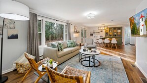 A modern living room with a sofa, coffee table, and chairs. There is a dining area with a table and chairs. The room has large windows and a view of trees outside. - Cozy, Quiet and Peaceful Bailey's Harbor home: Close to Everything Door County! (Baileys Harbor)