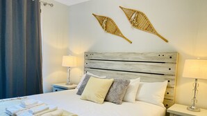 A neatly made bed with white linens, a beige throw blanket, and a headboard with a wooden finish. There are two bedside tables with lamps, and a curtain is partially visible on the left. - Spring Creek Luxury One Bedroom Sleeps 4 (Canmore)