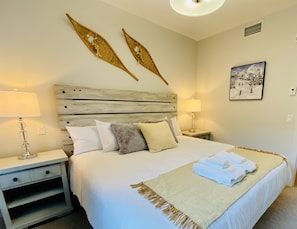 A neatly made bed with white linens and a beige throw blanket, flanked by two bedside tables with lamps, a wooden headboard, and a wall-mounted painting.