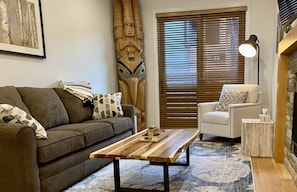 A living room with a wooden coffee table, a sofa, a chair, and a large window with blinds.