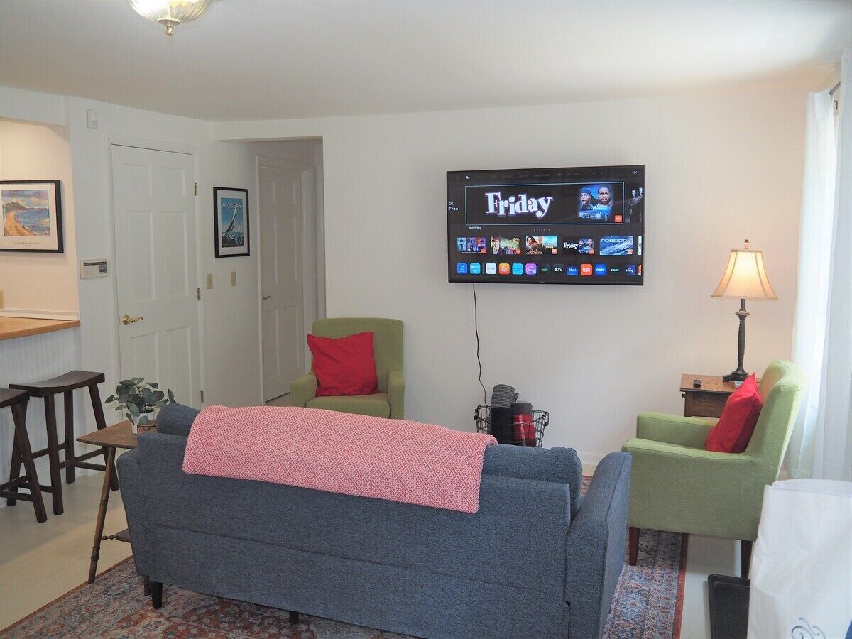 Ein Wohnzimmer mit grauem Sofa, zwei grünen Sesseln, einem an der Wand montierten Fernseher, der einen Freitagsprogramm anzeigt, und einem kleinen Tisch mit einer Lampe.