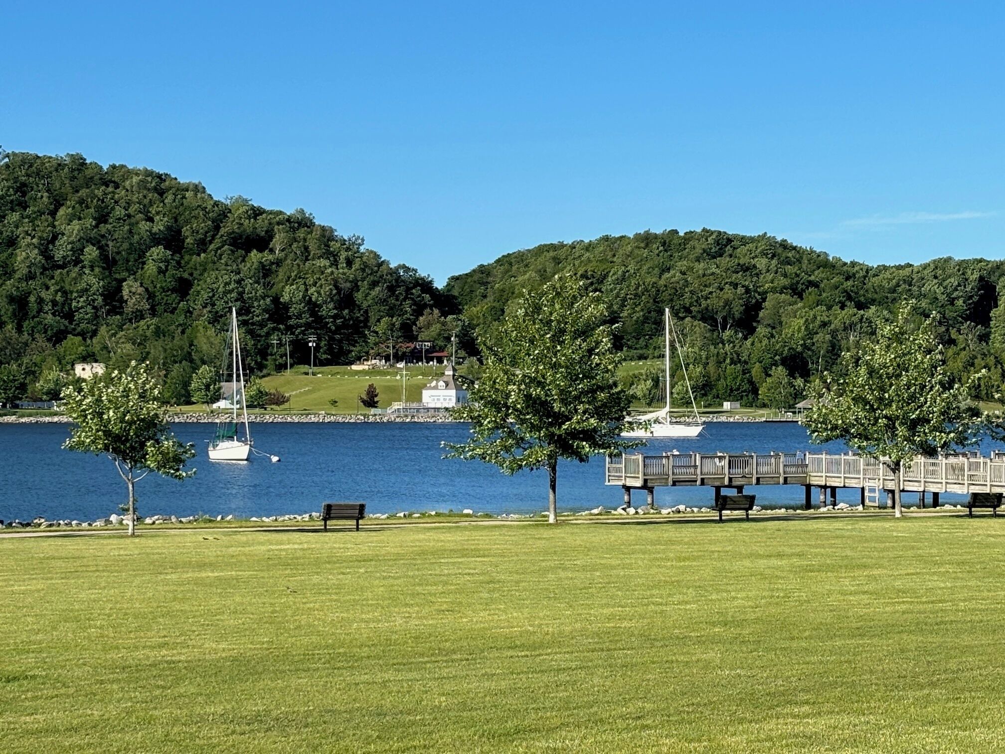 A lakeside park with a wooden pier, benches, and a sailboat.