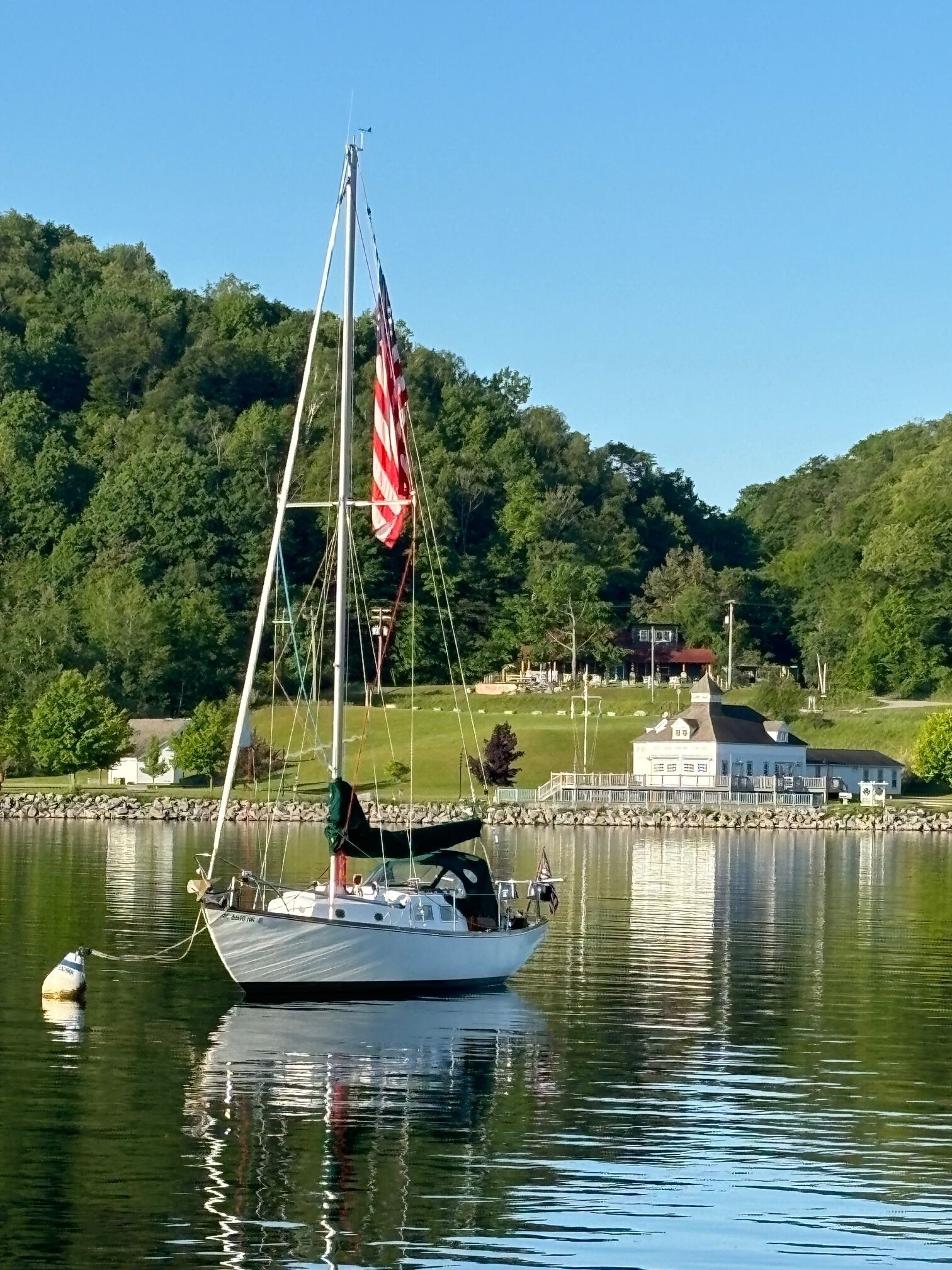 Un velero amarrado en un lago tranquilo con una colina arbolada al fondo y un edificio de fachada blanca.