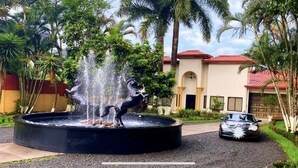 A fountain with a horse statue in a circular basin, surrounded by lush greenery and a building with a red-tiled roof in the background. - Quinta El Zable (Zaragoza)