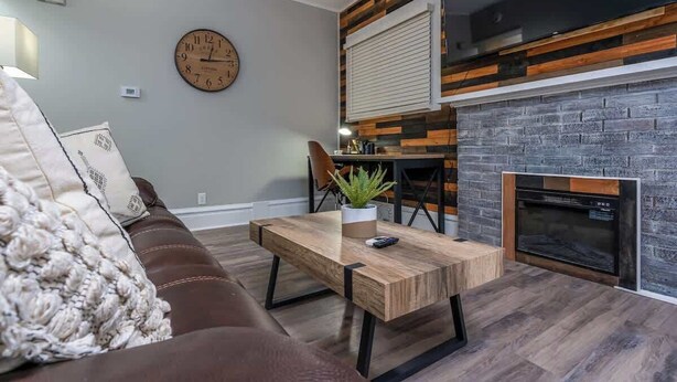 Een woonkamer met een bruine bank, een houten salontafel, een klok aan de muur en een open haard met een bakstenen gevel.