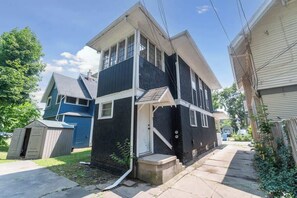 A two-story residential building with a front porch, a small attached garage, and a clear sky. - Remote Ready I 2 BR 1 BA (Lansing)