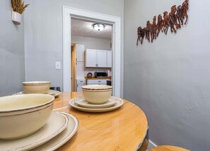 A dining area with a wooden table, white plates, and bowls. There is a kitchen visible in the background. - Remote Ready I 2 BR 1 BA (Lansing)