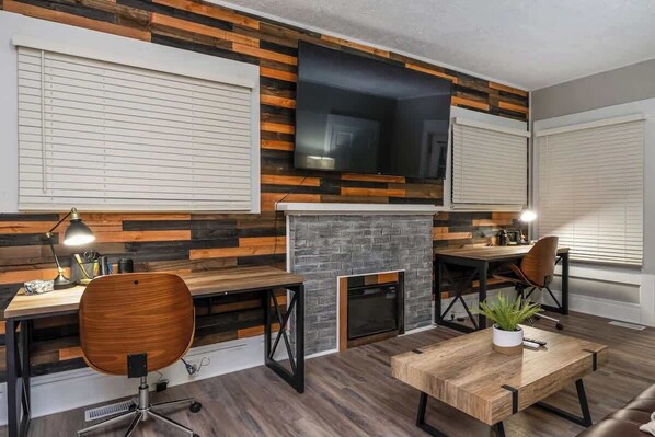A modern living room with a fireplace, wooden wall paneling, a flat-screen TV, and a dining area with a wooden table and chairs. - Remote Ready I 2 BR 1 BA (Lansing)