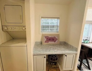 Une cuisine compacte dotée d’un lave-linge et d’un sèche-linge intégrés, d’une petite fenêtre avec des stores et d’un panneau indiquant « BAIGNADE INTERDITE » placé sur le plan de travail.