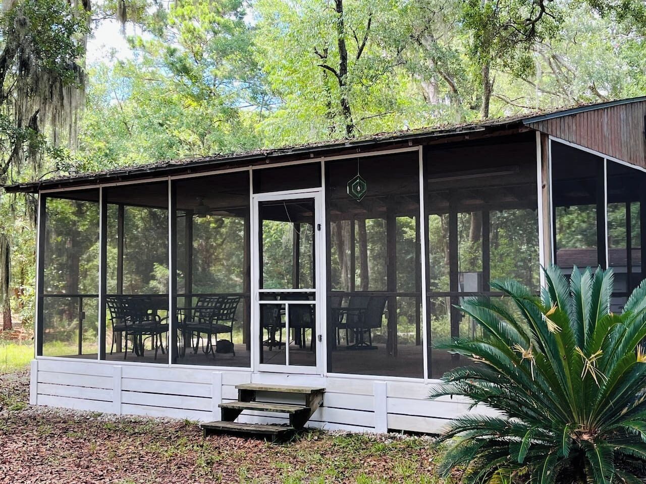 A covered outdoor seating area with a bench and chairs, surrounded by trees.