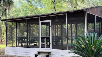 A covered outdoor seating area with a bench and chairs, surrounded by trees.