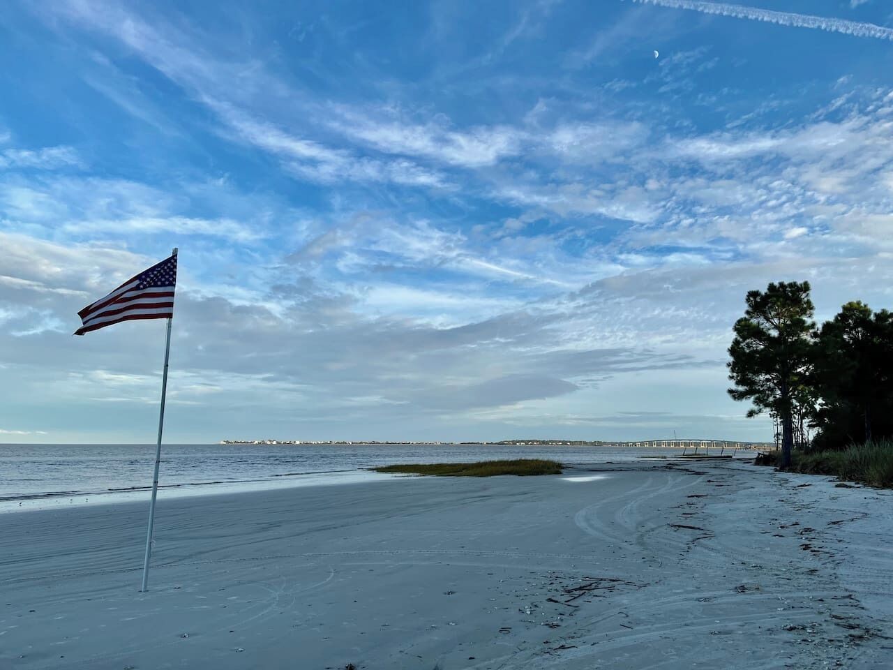 A beach with an American flag, a clear sky, and a distant bridge.