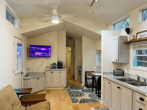 A compact kitchen with a TV mounted above the counter, a dining area with a table and chairs, and a ceiling fan. - Cute Cottage Next to Hunting Island. (Saint Helena Island)