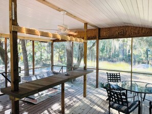 Terraza de madera al aire libre con una mesa y sillas, rodeada de grandes ventanales con vistas a árboles.