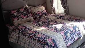 A bed with a floral-patterned bedspread and matching pillows, a tufted headboard, and a window with curtains. - Apartment Sleeps 4 with Balcony and Parking (Asaba)