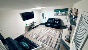 Una sala de estar con un sofá de cuero negro, un televisor montado en la pared, una mesita con un florero y un cuadro en la pared.