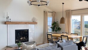 Ein modernes Wohnzimmer mit einer großen, grauen Eckcouch, einem Kamin, einem Essbereich mit Holztisch und Stühlen sowie Blick ins Freie durch große Fenster.