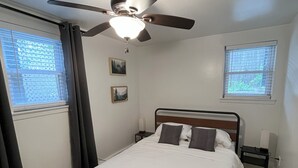 A bedroom with a bed, a ceiling fan, two windows with blinds, a nightstand, and a wall-mounted lamp. - Two Bedroom Private Apt near NYC! (West New York)