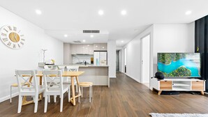 Una cocina moderna con armarios blancos, una mesa de comedor de madera y sillas. Hay un televisor de pantalla plana grande sobre un soporte blanco con base de madera.