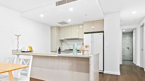 Una cocina moderna con armarios blancos, encimera de granito y horno empotrado. Hay una zona de comedor con mesa de madera y sillas blancas.