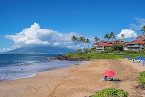 Una playa con una sombrilla roja, palmeras y un hotel con tejados rojos al fondo.