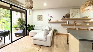A modern living room with a white sofa, a dining area with a dark countertop, and a view through large glass doors to an outdoor balcony. - Aloha Moon - The Honeymoon Hideaway (Wailea)