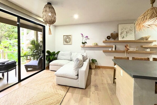 Una sala de estar moderna con un sofá blanco, un comedor con encimera oscura y vistas a un balcón exterior a través de amplios ventanales.