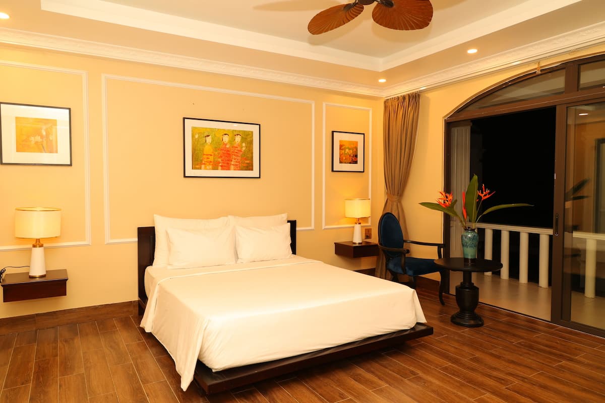A hotel room with a bed, a nightstand, a lamp, a chair, a vase with flowers, and a balcony with a railing.
