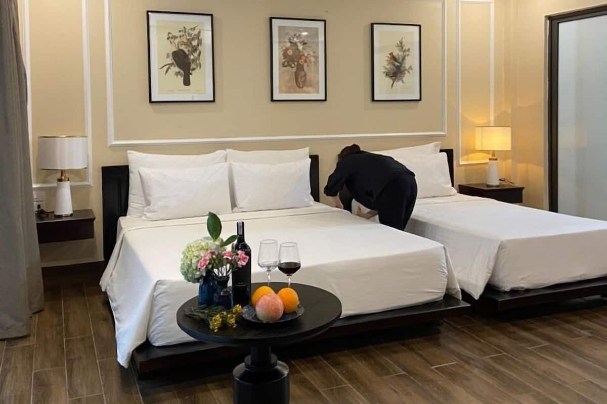 A hotel room with two beds, a ceiling fan, framed floral artwork on the wall, a small table with a bottle of wine and fruit, and a nightstand with a lamp.