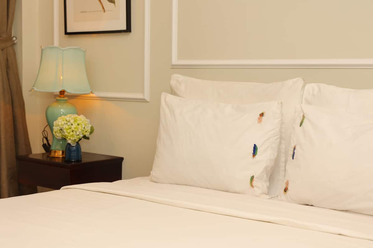 A neatly made bed with white linens and decorative pillows, a bedside table with a lamp and a vase of flowers, and a framed picture on the wall.