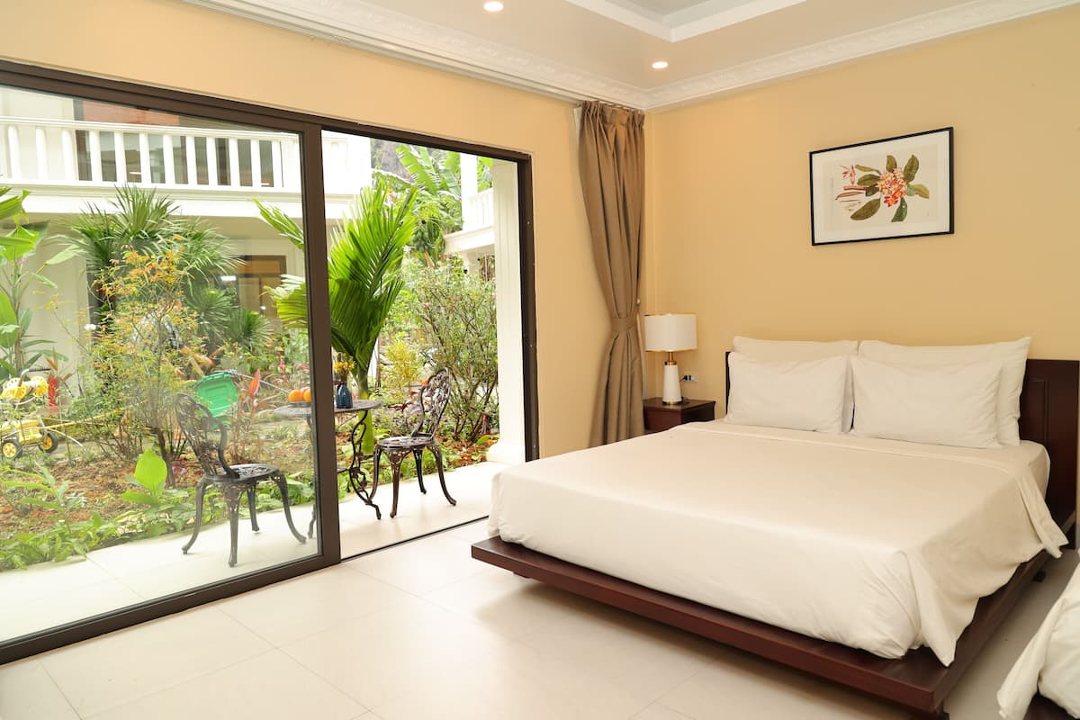 A bedroom with a large bed, a nightstand, a lamp, and a view of a garden through a sliding glass door.