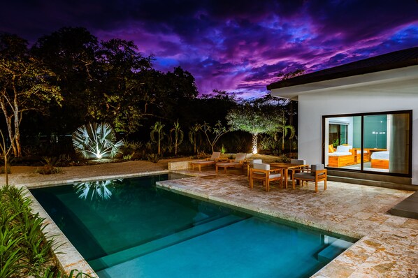 A modern outdoor pool area with a clear pool, wooden lounge chairs, and a well-lit patio.