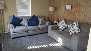 A living room with two grey sofas, each adorned with patterned cushions. A wooden wall panel with a framed inspirational quote is visible. - Quiet Space with Spacious Yard for Parking in Nice Community  (East Millsboro)