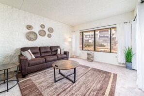 A living room with a brown leather sofa, a round coffee table, and a large window with curtains.